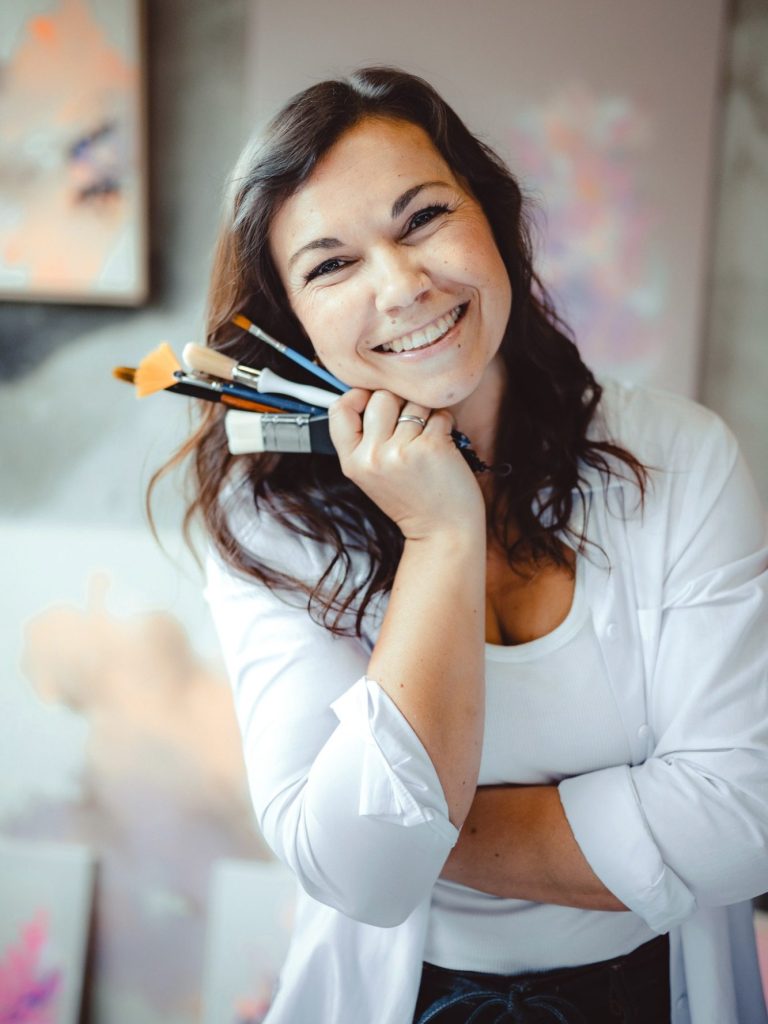 Künstlerin Anca Juliana Schulte hält Pinsel in der Hand, umgeben von Kunstwerken.