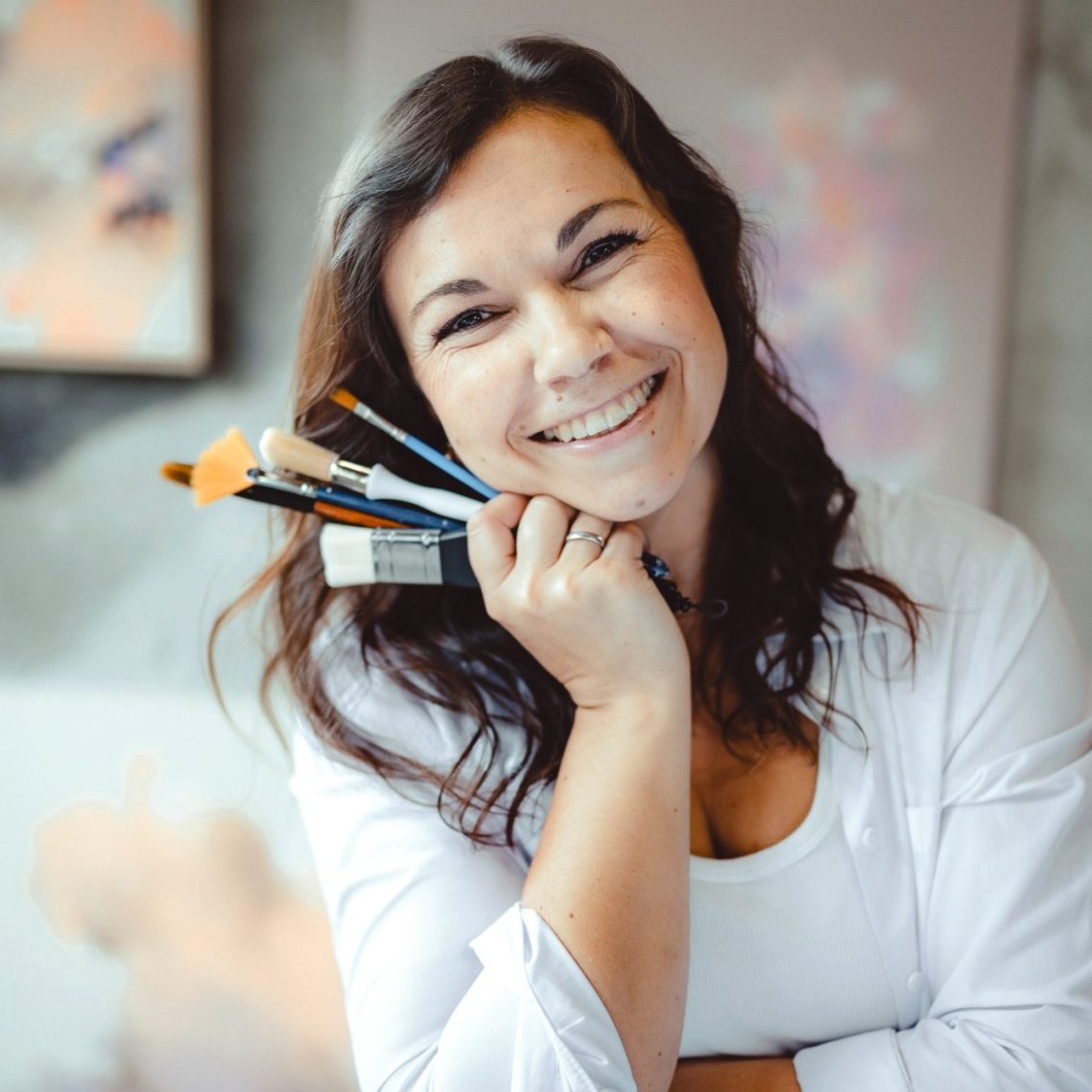 Lächelnde Anca Juliana Schulte hält Pinsel in der Hand, steht vor einem künstlerischen Hintergrund.