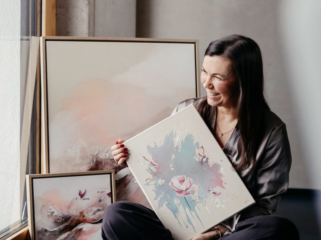 Anca Juliana Schulte sitzt lächelnd vor Kunstwerken in einem Atelier.