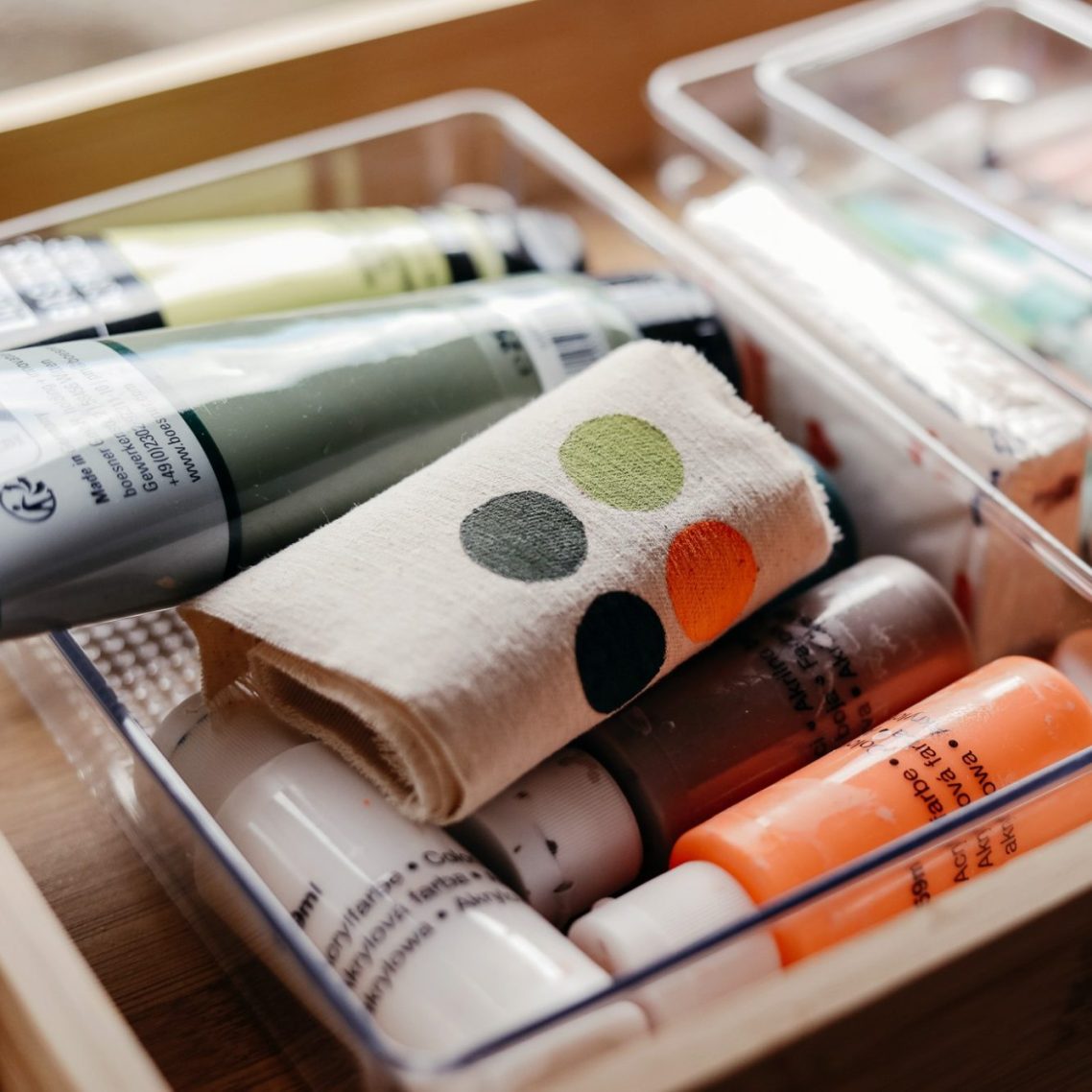 Farben und Malutensilien in einer transparenten Box, Stoffrolle mit Farbklecksen.