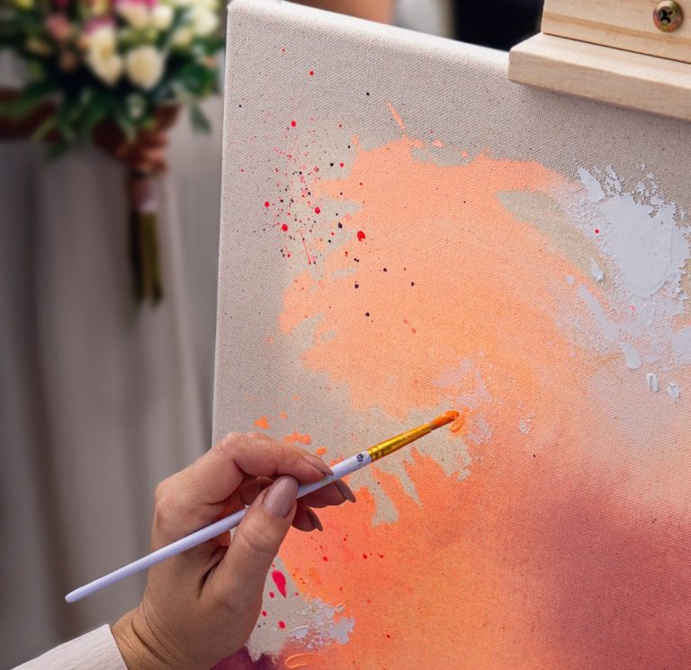 Eine Hand mit Pinsel malt orangefarbene und weiße Farbspritzer auf eine Leinwand.