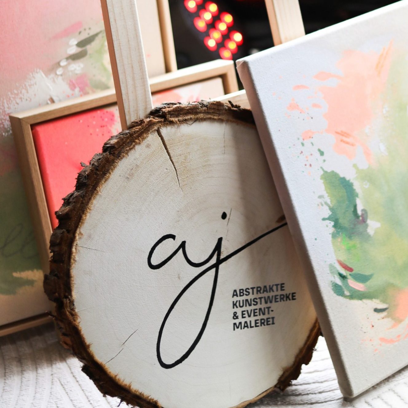 Holzscheibe mit Logo, umgeben von bunten, abstrakten Kunstwerken in Rahmung.