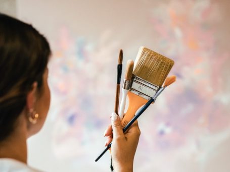 Anca Juliana hält Pinsel in der Hand vor einer pastellfarbenen Leinwand.